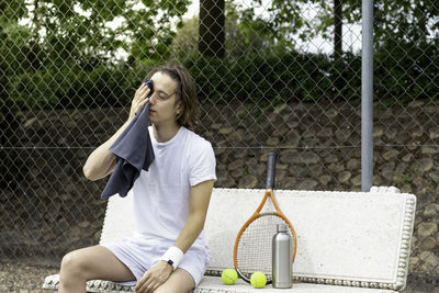 Young man wiping his sweat with a towel sitting on a bench after playing a game of tennis on a court