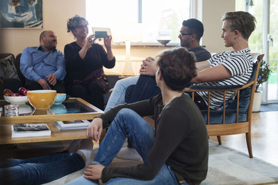 Smiling woman photographing friends while sitting by table in living room