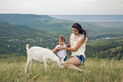 Woman with a child in a field on the mountain sitting next to a small goat in the summer