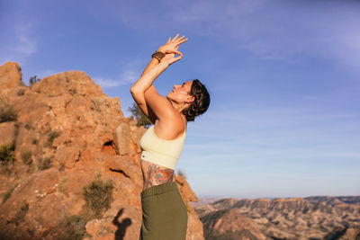 Side view of slim female in casual outfit performing mountain with arms up and backbend posture on slope of rocky mountain