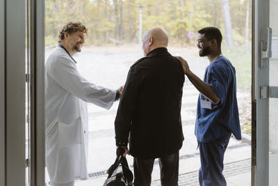 Senior male patient shaking hands with doctor while standing by nurse at hospital doorway