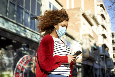 Curly hair woman wearing sanitary mask holding coffee cup while using mobile phone standing in city