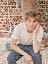 Portrait of young man talking over phone while sitting on swing against wall