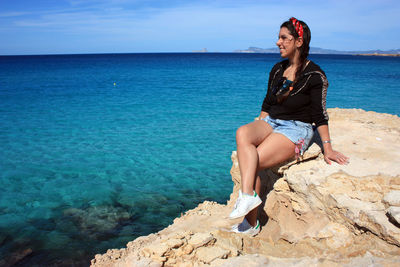 Young woman sitting on rock by sea against sky