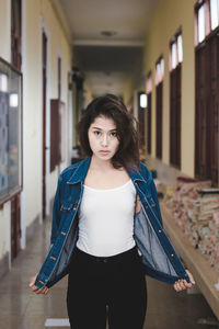 Portrait of young woman holding denim jacket while standing in corridor