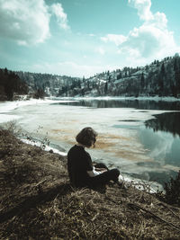 Side view of man sitting by lake against sky