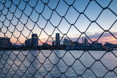 City seen through chainlink fence against sky during sunset