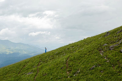 Woman in a blue long dress stands on top of a mountain in the summer on a green meadow