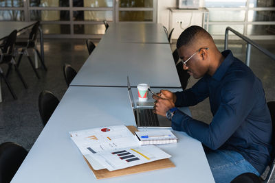 Side view of african american concentrated male in eyeglasses and formal clothes sitting in chair at table while using phone and netbook near document and takeaway drink in light workspace