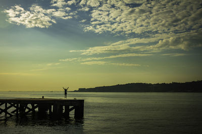 Silhouette of a man with arms outstretched while jumping off the pier during sunset. 