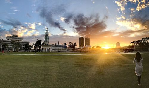 Rear view of woman walking on grass in city against sky during sunset