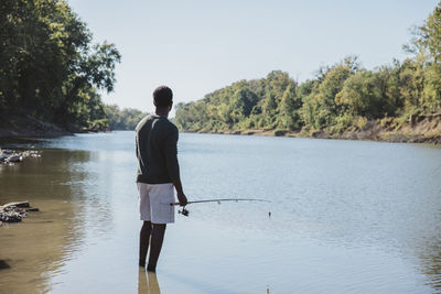 Rear view of man holding fishing rod while standing in lake against clear sky