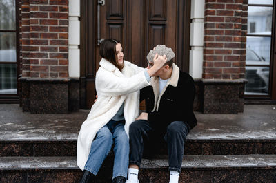  guy and a girl in fur coat are sitting on the porch of a brown brick house. 