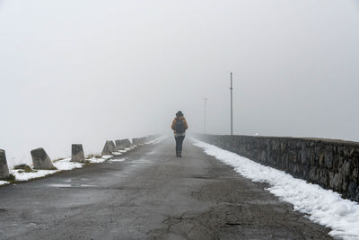 Rear view of person walking on road across a dam. foggy, moody, cold weather.