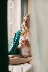 A young girl is sitting on the windowsill in the early morning, drinking hot coffee, 