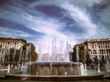 Fountain in city against cloudy sky