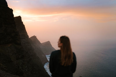 Woman standing on rock against sky during sunset