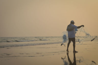 Rear view of man holding fishing net at beach against sky during sunset