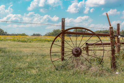 Rusty metallic wheel leaning on fence at field against sky