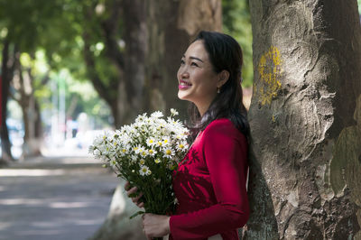 Side view of woman holding flower bouquet while standing by tree trunk