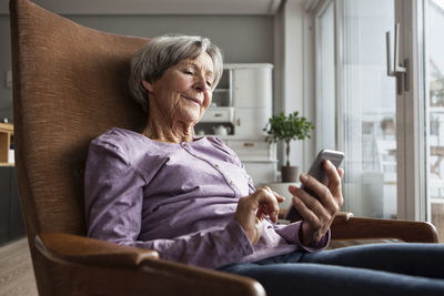 Portrait of senior woman sitting on armchair at home using smartphone