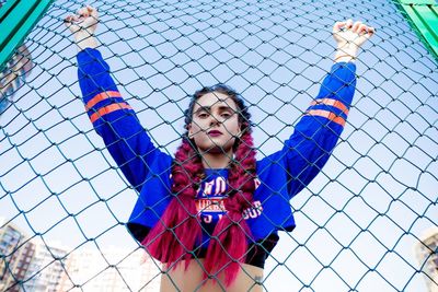 Portrait of young woman with arms raised standing by chainlink fence against sky