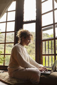 Young woman using mobile phone while sitting on window