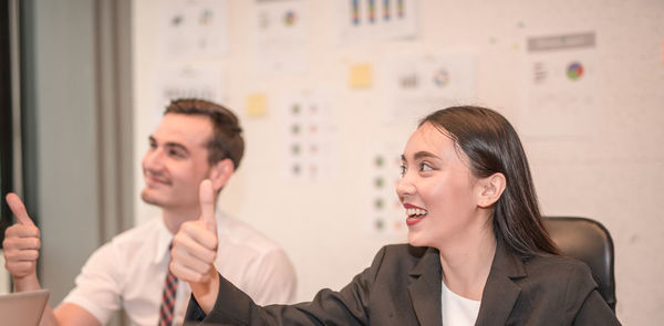 Smiling colleagues gesturing thumbs up sign while sitting in office
