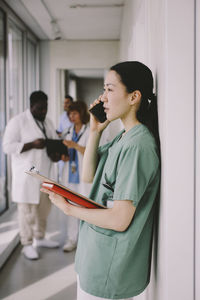 Side view of female doctor talking on smart phone while standing with clipboard in hospital corridor