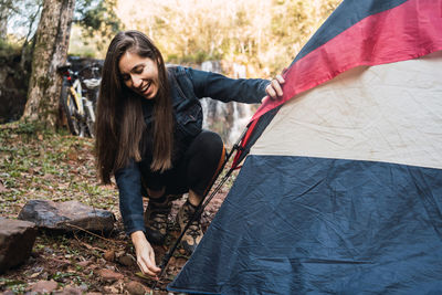 Full body of positive young female hiker sitting on haunches while setting up tent during weekend in woods