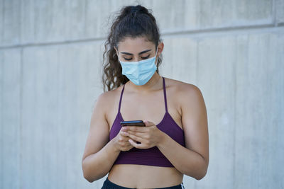 Young woman using phone while standing against wall