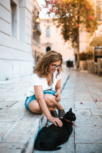 Smiling woman stroking cat on footpath in city