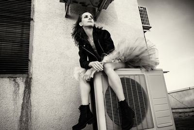 A ballerina in tutu, jacket and boots climbed on the air conditioner and poses against the sky