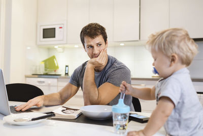 Father looking at baby boy while using laptop in kitchen
