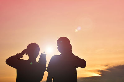 Silhouette girl with brother listening to music against sky during sunset