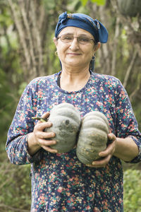 Portrait of smiling senior woman holding pumpkins against trees