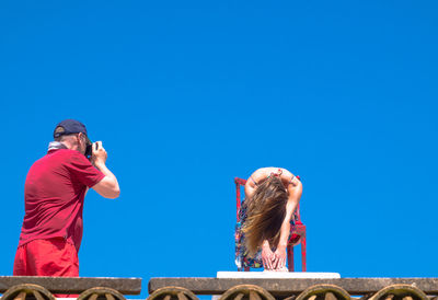 Man photographing woman sitting on chair against clear sky