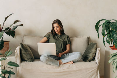 Young latin female student sitting on the sofa uses a laptop for online learning.