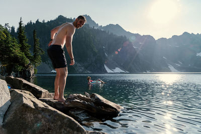 Portrait of shirtless mature man standing on rock at lakeshore against sky during sunny day