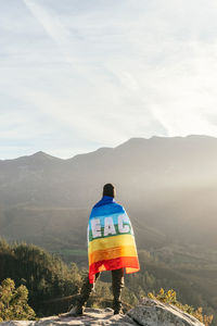 Back view of hiker standing with rainbow lgbt flag with inscription peace and enjoying freedom in mountains