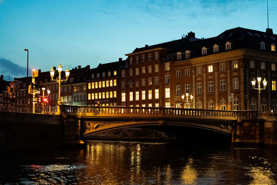 Bridge over river by illuminated buildings against sky at dusk