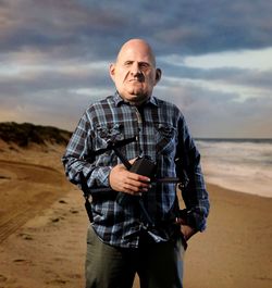 Portrait of mature man with camera standing at beach against cloudy sky