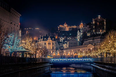 Illuminated bridge over river by buildings against sky at night