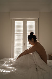 Side view of topless woman sitting on bed at home