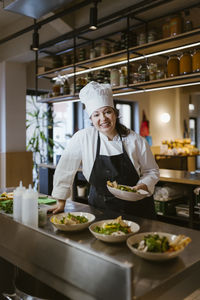 Smiling female chef holding salad bowl while leaning on kitchen counter at restaurant