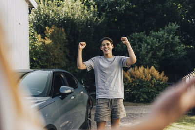 Cheerful young man holding fist while celebrating near car in back yard