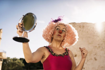 Confident mature female clown in bright costume and funny curly wig with makeup looking in round cosmetic mirror while standing on street on sunny day