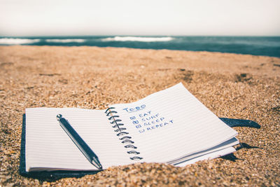 Close-up of spiral notebook with pen on sand at beach against sky