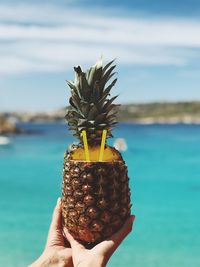 Cropped hands holding pineapple with drinking straws against sea