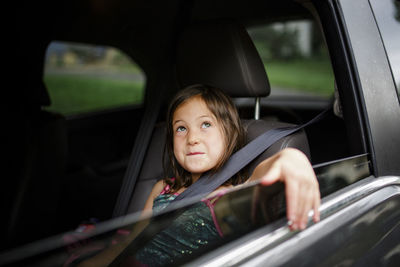 A little girl sits smiling in a car with her arm draped out the window